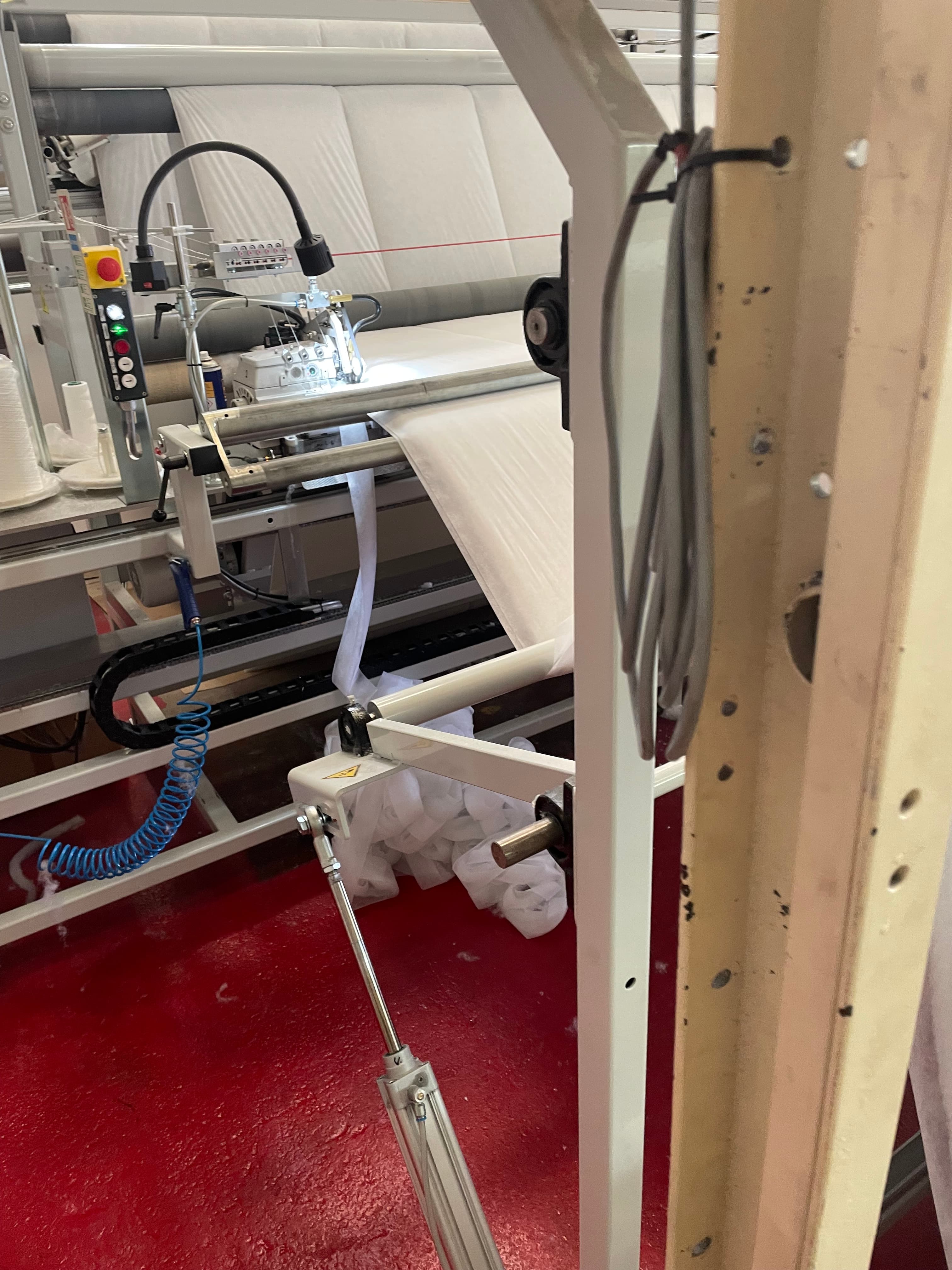 Quilting machine in a factory environment — thread cones on left, pneumatic components, red factory floor visible